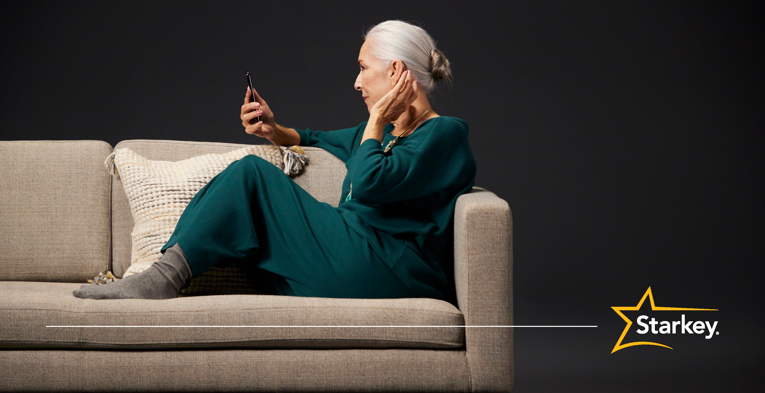 Senior woman sitting on couch holding a phone in right hand and left hand touching her left ear